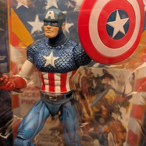 May include: Captain America action figure in a plastic package. The figure is wearing a blue helmet with a white "A", a blue and white scale-patterned top, and red, white, and blue striped torso. He holds a red, white, and blue shield.