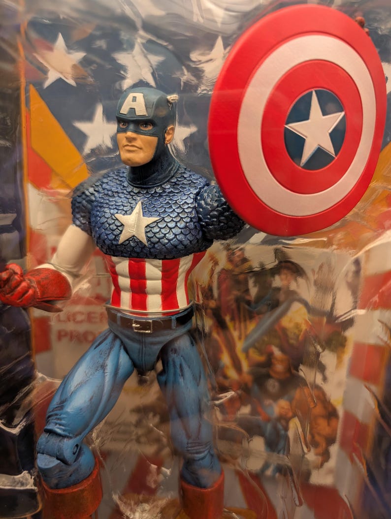 May include: Captain America action figure in a plastic package. The figure is wearing a blue helmet with a white "A", a blue and white scale-patterned top, and red, white, and blue striped torso. He holds a red, white, and blue shield.