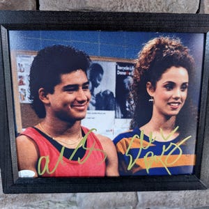 May include: Framed photograph featuring two people, a man and a woman, smiling. The man wears a red tank top, and the woman wears a blue and orange striped shirt. Both have autographs in green ink. The frame is black.