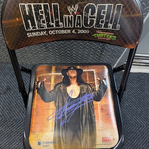May include: Folding chair featuring a wrestling theme. The backrest displays "Hell in a Cell" with the date "Sunday, October 4, 2009." The seat showcases a wrestler in a black coat with a signature. The chair has a black metal frame.
