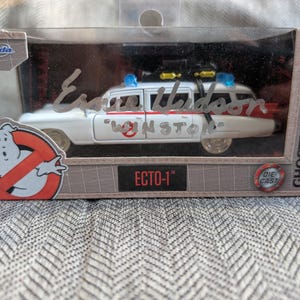 May include: A white die-cast model of the Ecto-1 from Ghostbusters, displayed in its original packaging. The car features red and white accents, blue lights, and the Ghostbusters logo. The packaging includes the text "ECTO-1" and "GHOSTBUSTERS".