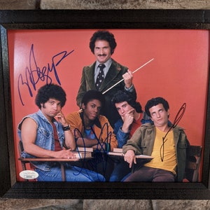 Puede incluir: Una foto enmarcada de cinco personas, cuatro hombres y una mujer, con sus firmas. La foto está sobre un fondo rojo. Las personas están todas sonriendo y parecen estar en una banda.