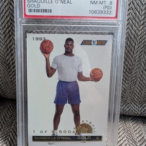 May include: A 1993 Classic Shaquille O'Neal Gold basketball card, graded NM-MT 8 by PSA, encased in a protective holder. The card features a photo of Shaquille O'Neal holding two basketballs. The text on the card reads "1 of 9,500".
