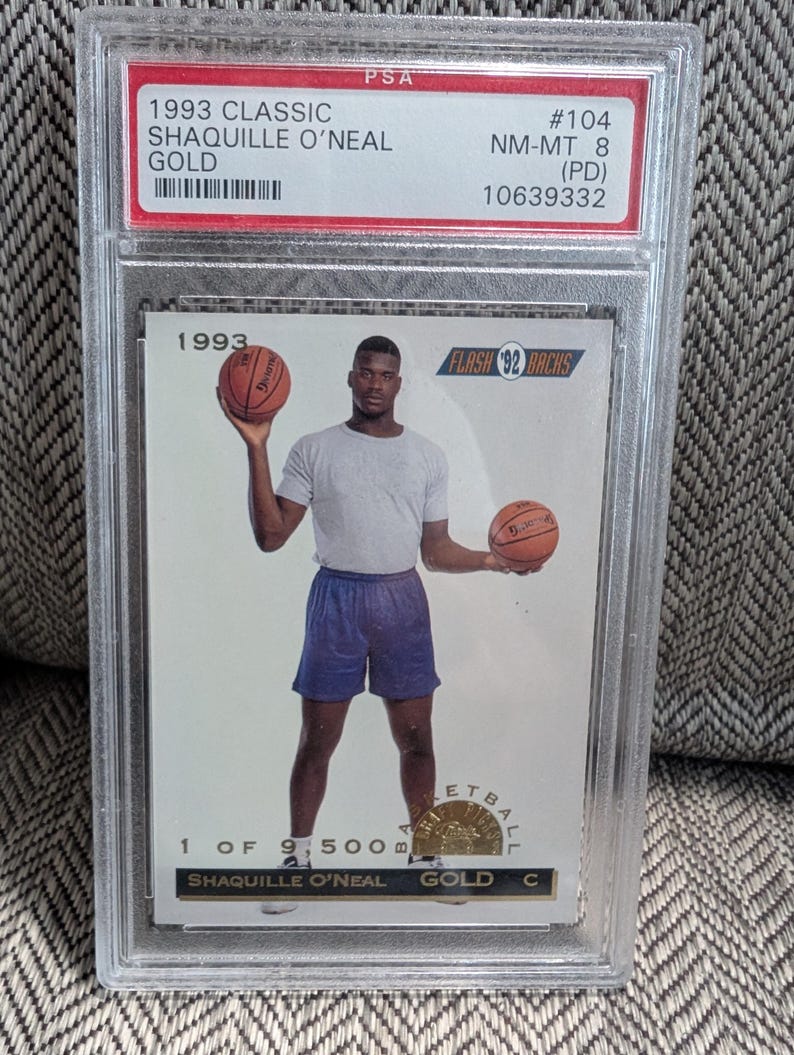 May include: A 1993 Classic Shaquille O'Neal Gold basketball card, graded NM-MT 8 by PSA, encased in a protective holder. The card features a photo of Shaquille O'Neal holding two basketballs. The text on the card reads "1 of 9,500".