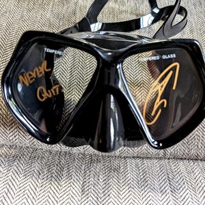 May include: Black diving mask with a wide frame and adjustable strap. The words "Never Quit" are written in gold on the left lens, and a gold signature is on the right lens. The mask is made of tempered glass.