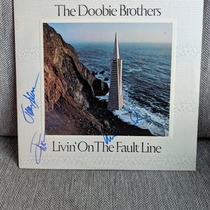 May include: Album cover for "Livin' On The Fault Line" by The Doobie Brothers. The cover features a photograph of a coastal landscape with a tall, pointed building. The album title and band name are printed on the cover, along with signatures.