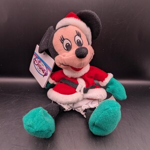 K&ouml;nnte beinhalten: Ein Minnie Mouse Pl&uuml;schtier in einem Weihnachtsmann-Outfit. Die Puppe hat einen roten Mantel mit wei&szlig;em Besatz, einen schwarzen G&uuml;rtel und einen passenden Weihnachtsmannhut. Gr&uuml;ne F&uuml;&szlig;e und H&auml;nde sind sichtbar. Ein Etikett von The Disney Store ist angebracht.