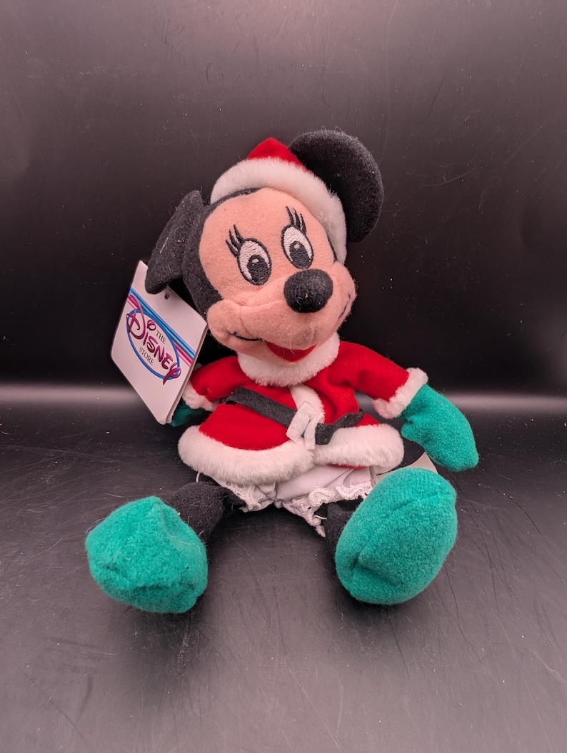 K&ouml;nnte beinhalten: Ein Minnie Mouse Pl&uuml;schtier in einem Weihnachtsmann-Outfit. Die Puppe hat einen roten Mantel mit wei&szlig;em Besatz, einen schwarzen G&uuml;rtel und einen passenden Weihnachtsmannhut. Gr&uuml;ne F&uuml;&szlig;e und H&auml;nde sind sichtbar. Ein Etikett von The Disney Store ist angebracht.
