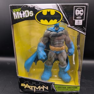 May include: A Batman action figure in a clear plastic display case. The figure is wearing a blue and gray costume with a yellow bat symbol. The packaging includes the text "Todd Mods," "DC Direct," and "Limited Edition Collector Vinyl."