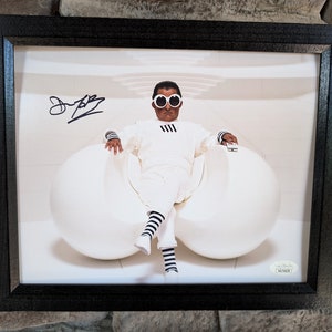 May include: A black and white photograph of a man wearing white clothing and large white sunglasses sitting in a white egg-shaped chair. The man is holding a small black object in his right hand. The photo is signed "Dr. Evil" in the top left corner.