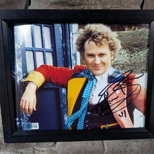 May include: Framed photograph featuring a man in a red jacket with yellow accents, a blue polka-dot tie, and a signed autograph. The image is set against a backdrop of a blue door and greenery, encased in a black frame.