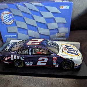 Op de afbeelding: Een modelauto van een NASCAR-raceauto, voornamelijk marineblauw en wit, met het nummer 2. De auto heeft sponsorlogo's en staat op een zwarte displaystandaard, met een blauwe doos op de achtergrond.