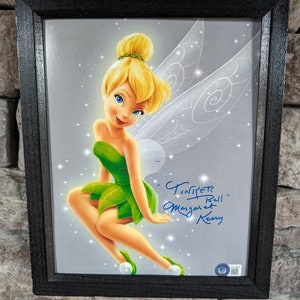 May include: A framed print of Tinker Bell, a Disney fairy with green wings and a green dress, sitting on a branch. The print is signed "Tinker Bell - Margaret Kerry".