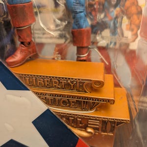 May include: Action figure of a superhero with blue and red costume. The figure is standing on a gold-colored platform with the words "LIBERTY JUSTICE ALL" engraved on it. The packaging includes a red, white, and blue design.