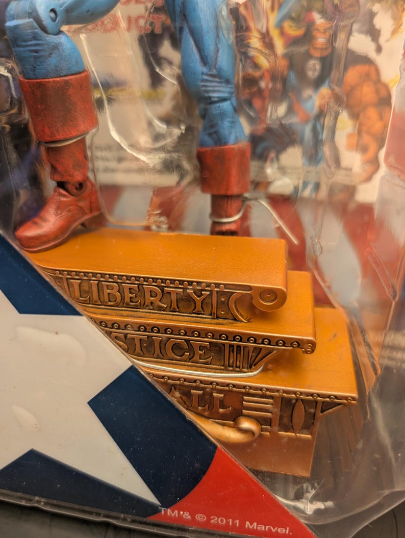 May include: Action figure of a superhero with blue and red costume. The figure is standing on a gold-colored platform with the words "LIBERTY JUSTICE ALL" engraved on it. The packaging includes a red, white, and blue design.