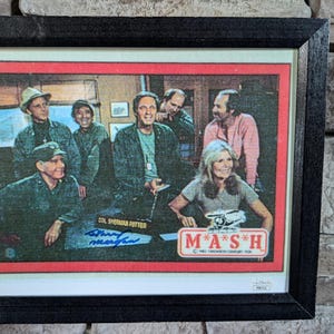 May include: A framed print of a vintage M*A*S*H television show trading card. The card features a group of actors from the show, including Alan Alda, Mike Farrell, and Loretta Swit. The card is signed by actor Harry Morgan, who played Colonel Sherman Potter on the show.