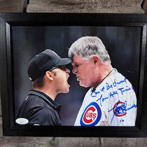 May include: A framed black and white photo of two men, one wearing a baseball uniform and the other wearing a black umpire uniform. The man in the uniform is signing the photo with the text "Can't Be Drew up Tommie Age 18 Time".