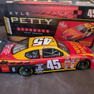 Op de afbeelding: Een model raceauto in rood en geel, met het nummer 45. De auto heeft sponsorlogo's en wordt geleverd met een doos met de afbeelding van de auto en de naam "Kyle Petty". De lengte is ongeveer 20 cm.