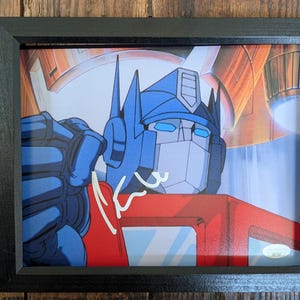 Op de afbeelding: Een ingelijste illustratie van het personage Optimus Prime met een blauw en rood kleurenschema, een metalen helm en gloeiende blauwe ogen. Het kunstwerk bevat een witte handgeschreven handtekening op de borst en een kleine authenticatiesticker.