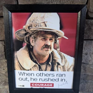 May include: Framed print featuring a firefighter covered in soot, wearing a helmet and coat. The print has the text "When others ran out, he rushed in. COURAGE" and a signature. The frame is black.