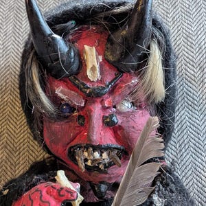 May include: A handmade doll with a red face, black horns, and white hair. The doll is wearing a black hooded cloak and holding a feather and a bone.