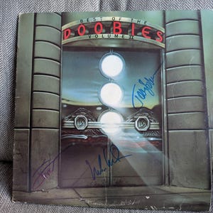 May include: Vintage vinyl record album cover for "Best of the Doobies Volume II" with signatures. The cover features a gray design with three white circles and the band's name in red. The album is on a gray surface.