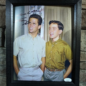 May include: A black framed photo of two young men, one with a patterned shirt and the other with a plaid shirt. The photo is signed "David Cameron" and "Bob" in black ink.