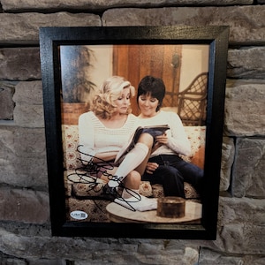 May include: Framed photograph of two women seated on a sofa, reading a book. The photo is signed with black ink. The frame is black and the photograph is mounted on a stone wall.
