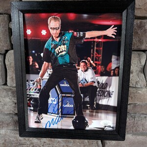 May include: A framed photograph of a man in a blue and green bowling shirt, throwing a bowling ball. The man is wearing sunglasses and has a determined expression on his face. The photo is signed by the man, and the signature is visible in the lower right corner of the image.