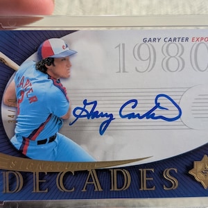 以下が含まれることがあります： 青いユニフォームを着たゲイリー・カーターの野球カード。テキスト「GARY CARTER EXPOS」と「1980s」が記載されています。カードにはサインと34/50の番号が記載されています。「DECADES」の文字が下部にあります。