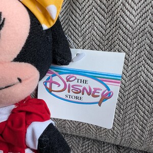 Puede incluir: Un peluche de Minnie Mouse con un lazo amarillo con lunares y un vestido rojo con lunares blancos. La etiqueta de Disney Store es visible.