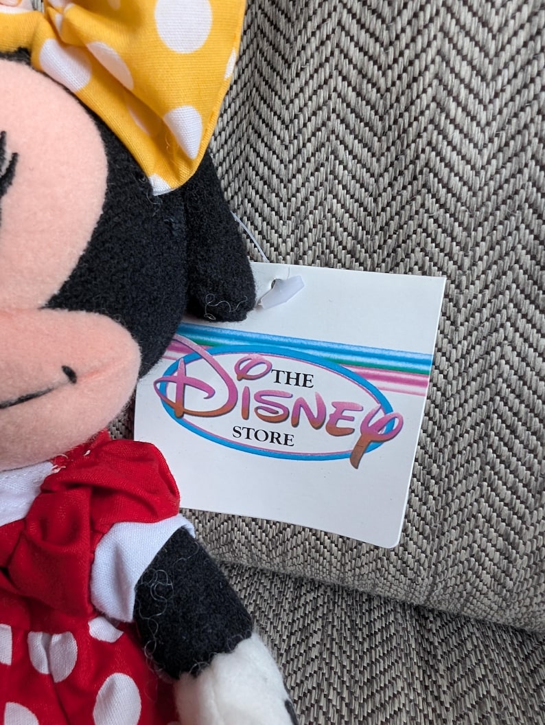 Puede incluir: Un peluche de Minnie Mouse con un lazo amarillo con lunares y un vestido rojo con lunares blancos. La etiqueta de Disney Store es visible.