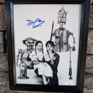 May include: Black and white photograph of a man, a girl, and two robots. The man is holding the girl on his lap. The girl is wearing a dress and has pigtails. The robots are made of metal and have human-like features. The photograph is signed "D.B."