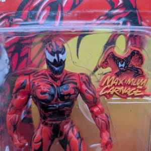 May include: A red and black action figure of the character Carnage from the Marvel Comics series. The figure is in a plastic package with the words "Maximum Carnage" and a bonus collector pin.