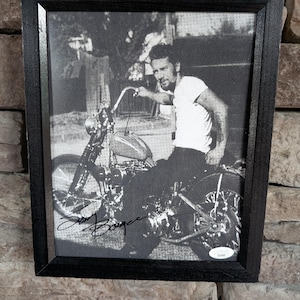 Puede incluir: Fotografía enmarcada en blanco y negro de un hombre en una motocicleta vintage. La fotografía está firmada y presenta un marco de madera negro. El hombre lleva una camiseta blanca y está sentado en la motocicleta.