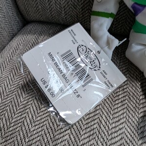 Op de afbeelding: Een Disney Mini Bean Bag Buzz 20 cm pop met een label. Het label is wit met zwarte tekst en een barcode. De pop is gedeeltelijk zichtbaar, met witte, groene en paarse accenten.