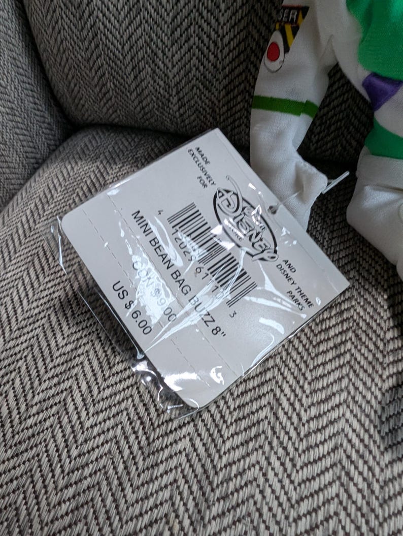 Op de afbeelding: Een Disney Mini Bean Bag Buzz 20 cm pop met een label. Het label is wit met zwarte tekst en een barcode. De pop is gedeeltelijk zichtbaar, met witte, groene en paarse accenten.