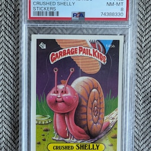 Könnte beinhalten: Eine Garbage Pail Kids Sammelkarte mit einer rosa Schnecke mit einer lila und orangefarbenen Schale. Die Karte trägt den Titel "Crushed Shelly" und die Nummer #145b. Die Karte ist von PSA mit NM-MT 8 bewertet.