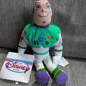 Op de afbeelding: Een Buzz Lightyear pluche pop van The Disney Store. De pop heeft een groen en wit ruimtepak met een Lightyear-patch, paarse details en een lachend gezicht. Een Disney Store-label is bevestigd.