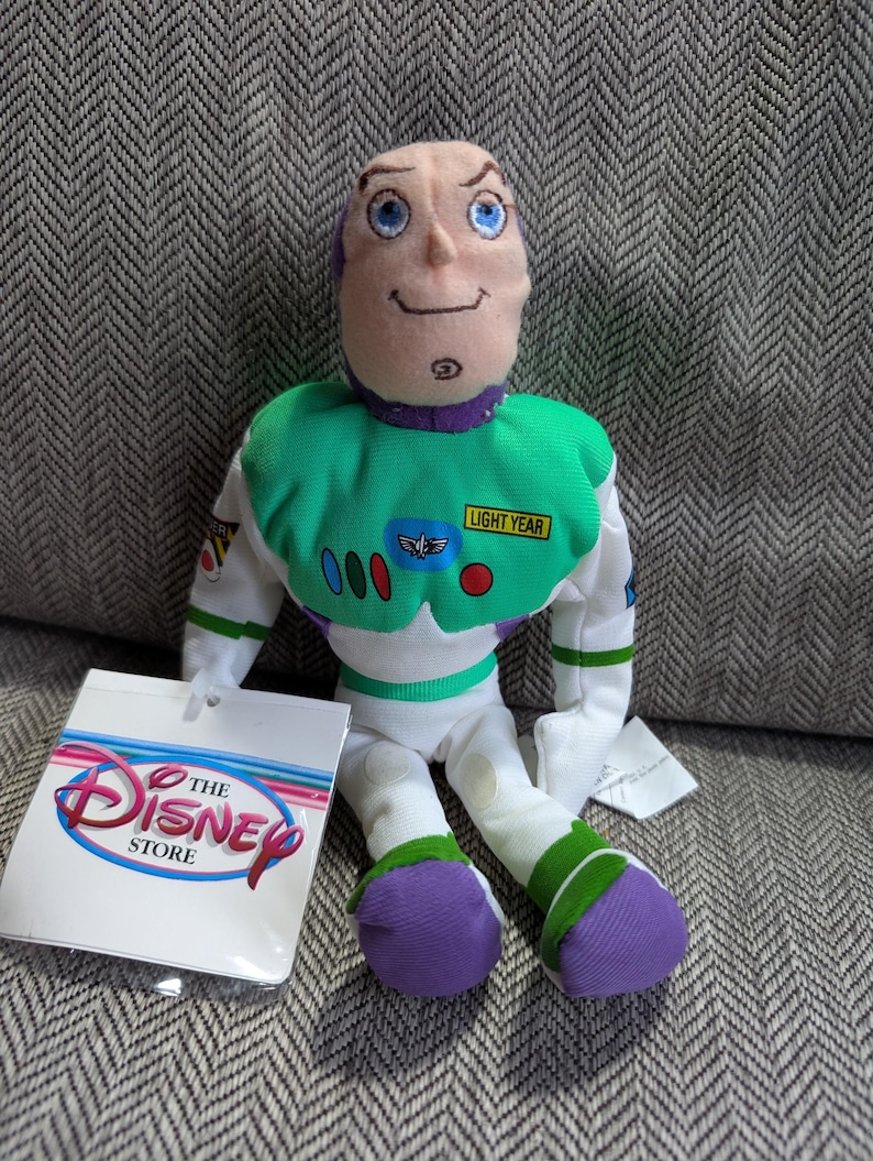 Op de afbeelding: Een Buzz Lightyear pluche pop van The Disney Store. De pop heeft een groen en wit ruimtepak met een Lightyear-patch, paarse details en een lachend gezicht. Een Disney Store-label is bevestigd.