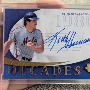 以下が含まれることがあります： ニューヨーク・メッツのキース・ヘルナンデスをフィーチャーした野球カード。カードは「Signature Decades」と「1980s」の文字が入った青と金のデザインです。カードは透明な保護ホルダーに入っています。