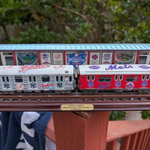 May include: Miniature subway train set featuring a Yankees train and a Mets train, with a station backdrop. The Yankees train is white with blue accents, and the Mets train is red with blue accents. The set is displayed on a wooden base.
