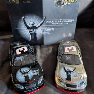 Op de afbeelding: Twee modelraceauto's, één zwart en één zilver, met het nummer 3 op het dak. De auto's hebben het logo van de Dale Earnhardt Foundation en de tekst "Monte Carlo". Een doos met het logo Race Fans Collectibles staat op de achtergrond.