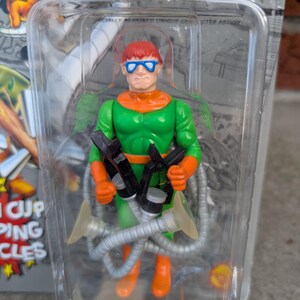 May include: Dr. Octopus action figure in a clear plastic package. The figure is green, orange, and red, with blue glasses and four mechanical arms. The packaging has the words "SUPER VILLAINS" and "DR. OCTOPUS" printed on it.