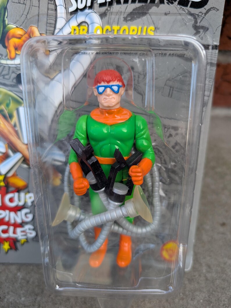 May include: Dr. Octopus action figure in a clear plastic package. The figure is green, orange, and red, with blue glasses and four mechanical arms. The packaging has the words "SUPER VILLAINS" and "DR. OCTOPUS" printed on it.