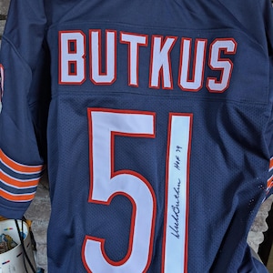 Puede incluir: Camiseta de fútbol azul marino con el nombre "BUTKUS" en letras blancas con ribete naranja en los hombros. El número "51" está en la parte delantera y trasera en blanco con ribete naranja. Incluye una firma y "HOF 79".