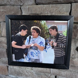 May include: Framed photograph featuring three men, each with autographs. The image shows the men in casual attire, with one wearing a striped shirt and another wearing a black t-shirt. The frame is black and the background is a stone wall.