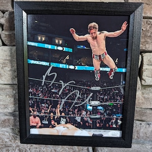 Könnte beinhalten: Ein schwarz-weiß gerahmtes Foto eines Wrestlers in der Luft, der einen Move in einem Wrestling-Ring ausführt. Der Wrestler trägt ein rot-weiß-blaues Wrestling-Outfit. Das Foto ist vom Wrestler signiert.