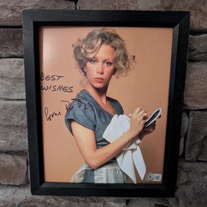 以下が含まれることがあります： ブロンドの髪の女性が、青いトップスを着て、白い布と小さなメモ帳を持っている額入りの写真。黒いフレームが画像を囲み、「Best Wishes」のサインが入っています。背景は明るい茶色です。