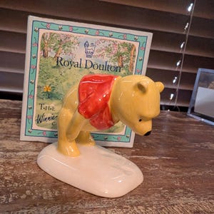 Puede incluir: Una figurita de cerámica amarilla de Winnie the Pooh con una camisa roja. La figurita está de pie sobre una base blanca y sostiene un tarro de miel. La figurita está delante de una caja con las palabras "Royal Doulton" y "The Winnie" en ella.